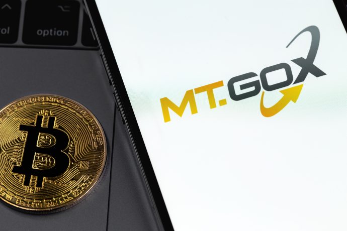 Eine Bitcoin-Münze liegt neben einem Smartphone. Auf ihrem Bildschirm leuchtet das ikonische MT Gox-Logo und hält einen Moment in der Geschichte der Kryptowährung fest.