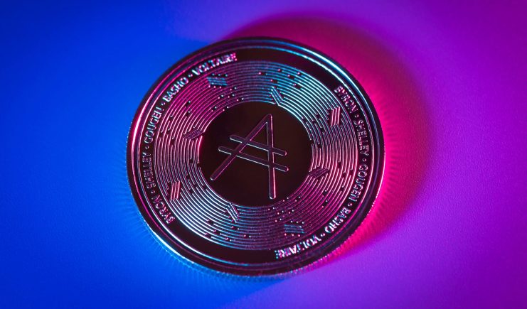 Eine Cardano (ADA)-Münze auf einem Farbverlaufshintergrund mit blauer und rosa Beleuchtung, mit dem Cardano-Logo