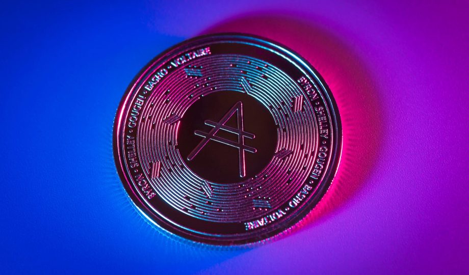 Eine Cardano (ADA)-Münze auf einem Farbverlaufshintergrund mit blauer und rosa Beleuchtung, mit dem Cardano-Logo