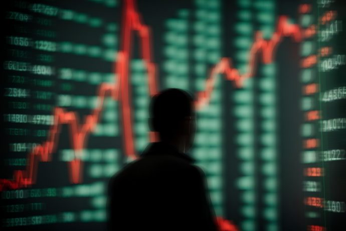 Silhouette einer Person vor einer digitalen Finanzanzeige mit roten und grünen Zahlen, die einer Schallwelle schwankender Diagramme ähnelt.