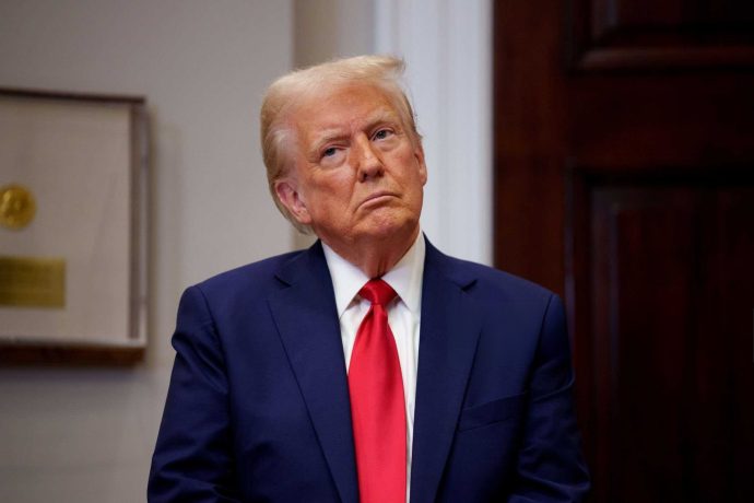 Eine Person in marineblauem Anzug und roter Krawatte steht im Innenbereich und blickt mit neutralem Gesichtsausdruck nach oben, der an Donald Trumps ikonische Posen erinnert. Im Hintergrund sind ein gerahmter Gegenstand und eine Holztür zu sehen, die ein Gefühl von Autorität und Bedächtigkeit vermitteln.