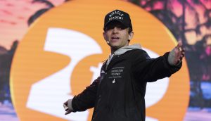 Eine Person in einer schwarzen Jacke und einer Miracle Academy-Kappe gestikuliert mit ausgebreiteten Armen auf der Bühne vor einem großen orange-weißen Hintergrund mit Palmenmuster und erinnert damit an die dynamische Präsenz von Jack Mallers bei einem Twenty One- oder MicroStrategy-Event.