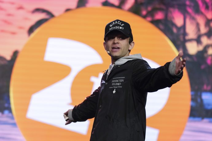 Eine Person in einer schwarzen Jacke und einer Miracle Academy-Kappe gestikuliert mit ausgebreiteten Armen auf der Bühne vor einem großen orange-weißen Hintergrund mit Palmenmuster und erinnert damit an die dynamische Präsenz von Jack Mallers bei einem Twenty One- oder MicroStrategy-Event.