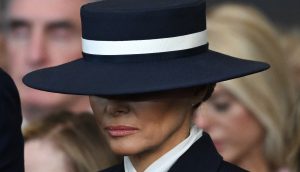 Melania Trump trägt einen breitkrempigen marineblauen Hut mit einem weißen Band, das ihr Gesicht teilweise verdeckt, und einen dunklen Anzug mit weißem Kragen.