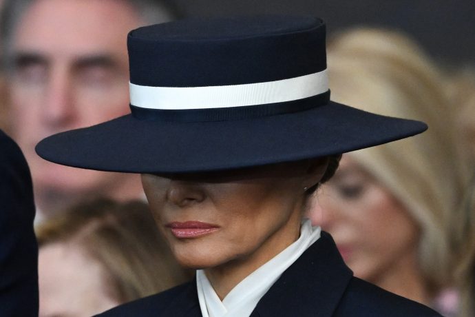Melania Trump trägt einen breitkrempigen marineblauen Hut mit einem weißen Band, das ihr Gesicht teilweise verdeckt, und einen dunklen Anzug mit weißem Kragen.