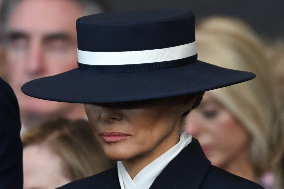 Melania Trump trägt einen breitkrempigen marineblauen Hut mit einem weißen Band, das ihr Gesicht teilweise verdeckt, und einen dunklen Anzug mit weißem Kragen.