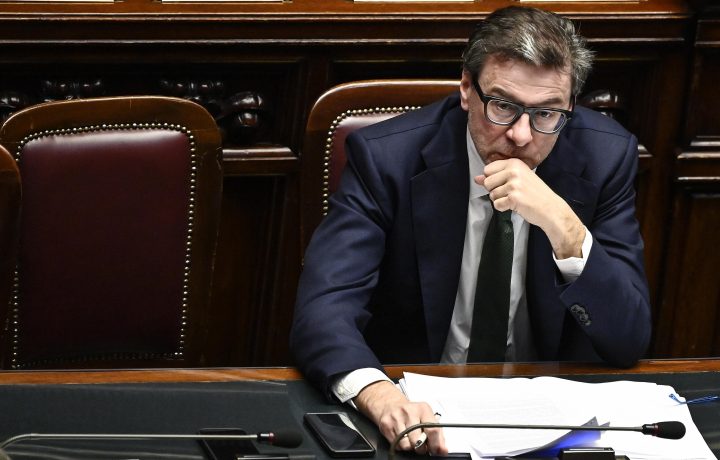 Ein Mann in dunklem Anzug und Brille, der einem italienischen Finanzminister ähnelt, sitzt an einem Schreibtisch mit Dokumenten, einem Telefon und einem Mikrofon und wirkt in einem formellen Umfeld mit Holzstühlen nachdenklich.