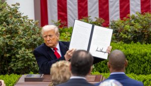 Donald Trump ein unterzeichnetes Dokument zur Feier des Befreiungstages hoch. Er steht auf einem Podium im Freien, im Hintergrund ist eine amerikanische Flagge zu sehen, und mehrere Personen beobachten ihn.