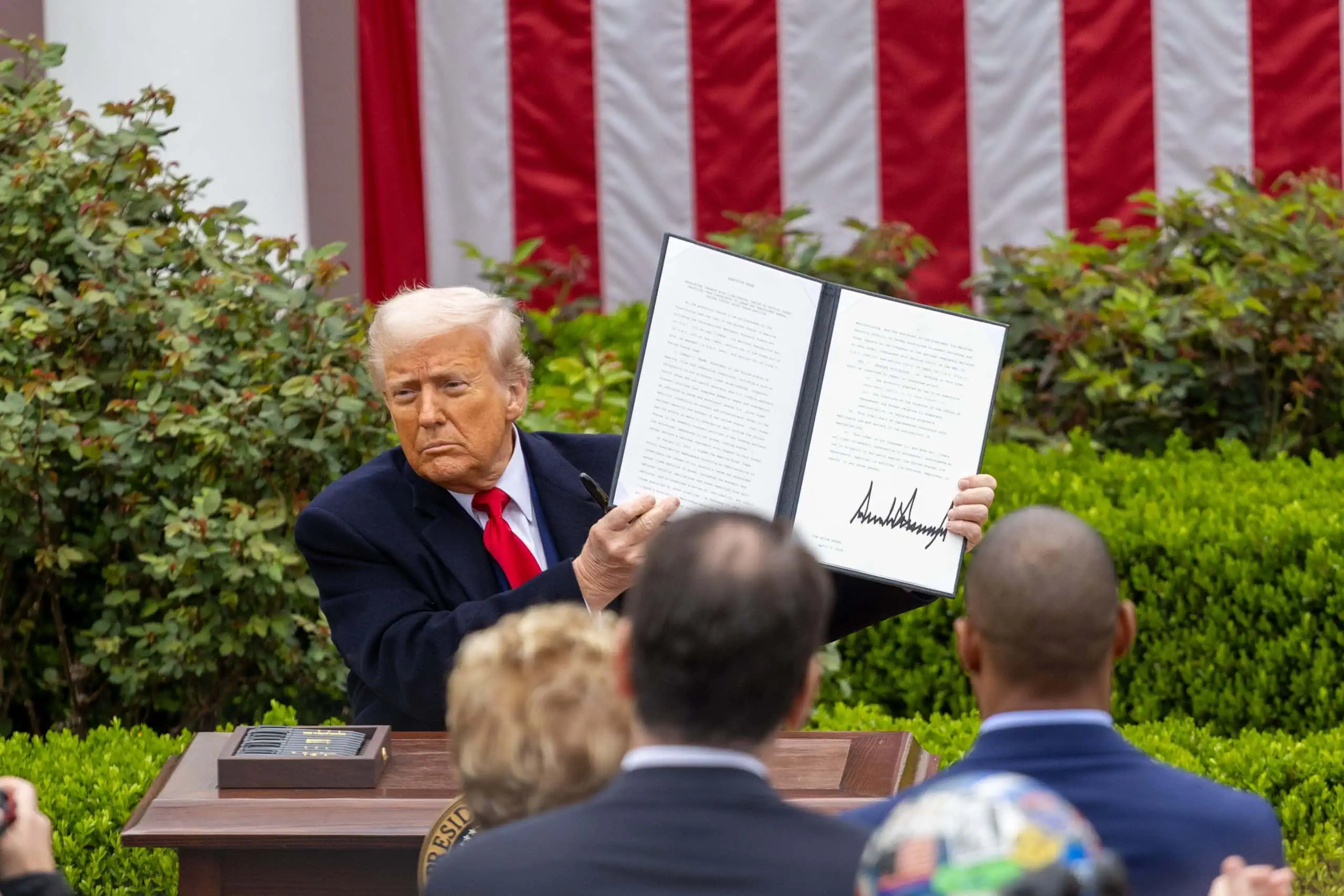 Donald Trump ein unterzeichnetes Dokument zur Feier des Befreiungstages hoch. Er steht auf einem Podium im Freien, im Hintergrund ist eine amerikanische Flagge zu sehen, und mehrere Personen beobachten ihn.