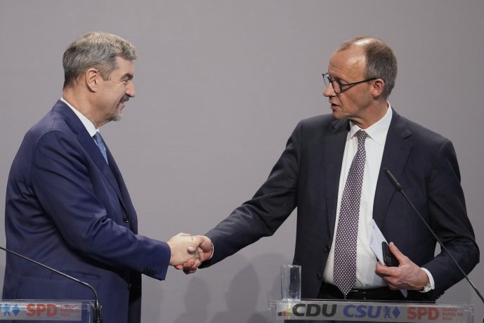 Zwei Männer in Anzügen geben sich auf der Bühne die Hand. Einer hält Papiere und einen Stift in der Hand – möglicherweise ein Zeichen für den Beginn einer neuen Legislaturperiode. Auf den Podesten prangen stolz die Logos von CDU, CSU und SPD vor grauem Hintergrund.