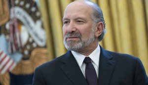 Howard Lutnick, US-Handelsminister, in Anzug und Krawatte steht vor einem unscharfen Hintergrund, auf dem ein Teil einer US-Flagge und ein Präsidentensiegel zu sehen sind.