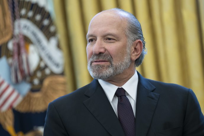 Howard Lutnick, US-Handelsminister, in Anzug und Krawatte steht vor einem unscharfen Hintergrund, auf dem ein Teil einer US-Flagge und ein Präsidentensiegel zu sehen sind.