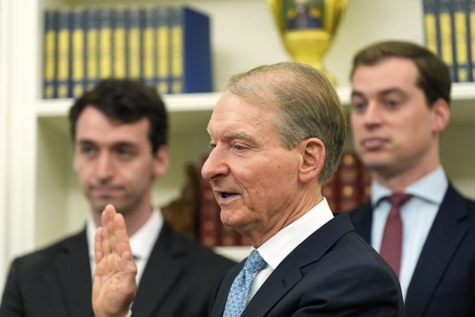 SEC-Vorsitzender Paul Atkins hebt enthusiastisch seine rechte Hand zur Amtseinführung, flankiert von zwei weiteren Männern in Anzügen, während Bücherregale und eine Trophäe den Hintergrund hervorheben.