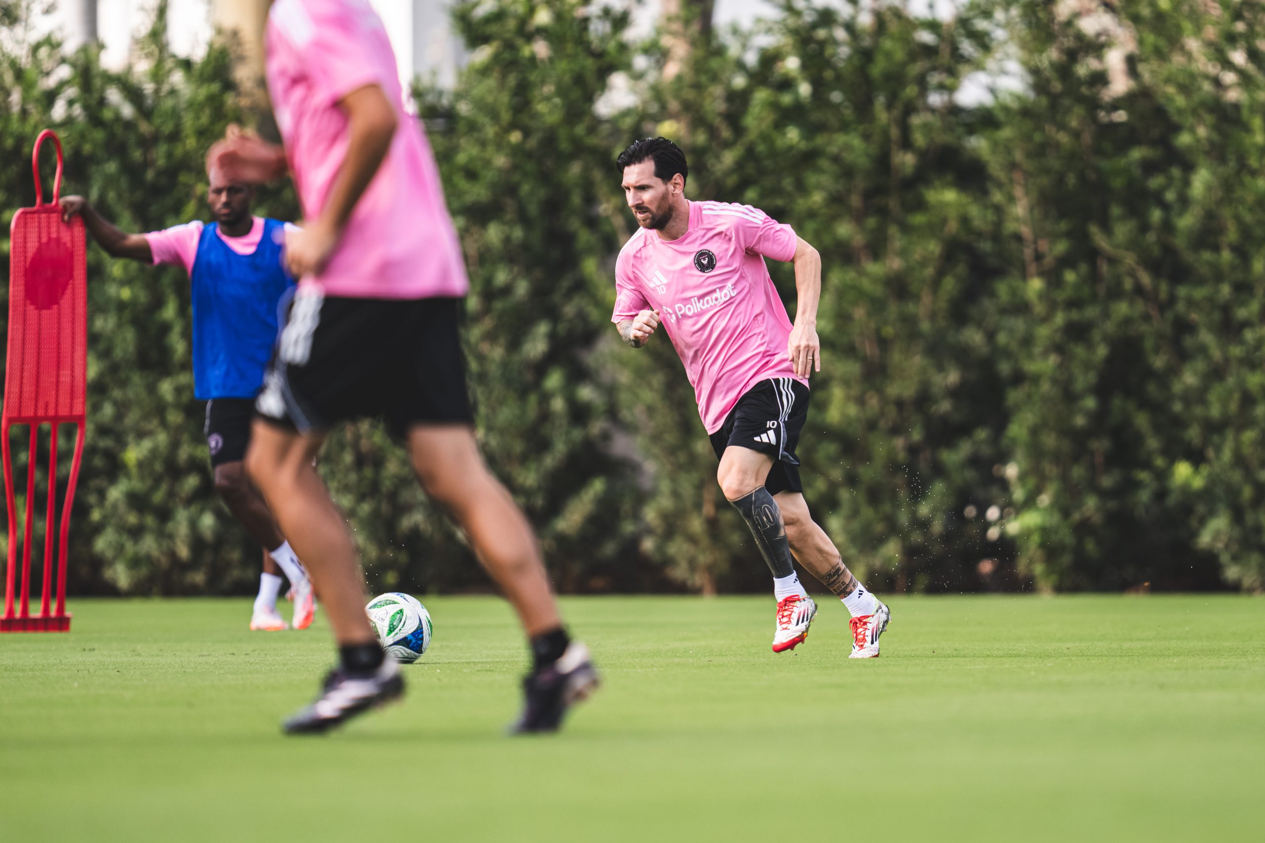 Ein Fußballspieler in einem rosa Trikot, das mit dezenten Tupfen verziert ist, dribbelt beim Training gekonnt einen Ball auf dem Rasenplatz, während seine Teamkollegen und eine Reihe von Ausrüstungsgegenständen den Hintergrund füllen.