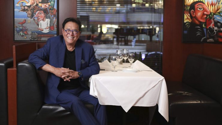 Ein Mann im blauen Anzug sitzt an einem Tisch in einem Restaurant mit schwarzen Sitznischen, weißen Tischdecken und farbenfrohen Kunstwerken und diskutiert möglicherweise über Robert Kiyosakis neueste Bitcoin-Prognose.