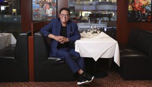 Ein Mann im blauen Anzug sitzt lächelnd an einem Tisch in einer Restaurantnische, hinter ihm hängen bunte Gemälde an der Wand – vielleicht diskutiert er über BTC oder Robert Kiyosakis Vorhersage, dass Bitcoin die Marke von 1.000.000 US-Dollar erreichen wird.