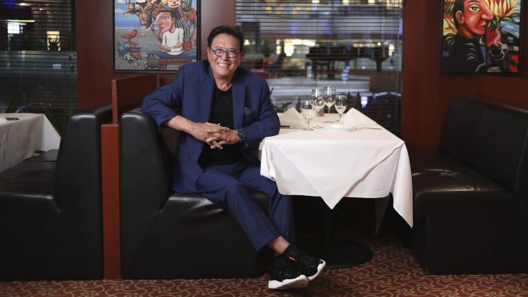 Ein Mann im blauen Anzug sitzt lächelnd an einem Tisch in einer Restaurantnische, hinter ihm hängen bunte Gemälde an der Wand – vielleicht diskutiert er über BTC oder Robert Kiyosakis Vorhersage, dass Bitcoin die Marke von 1.000.000 US-Dollar erreichen wird.