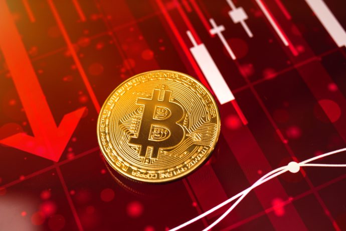 Eine goldene Bitcoin-Münze auf einem roten Finanzdiagramm-Hintergrund mit Abwärtspfeilen und Kerzenmustern, die die Auswirkungen von Trumps Zolloffensive widerspiegeln.