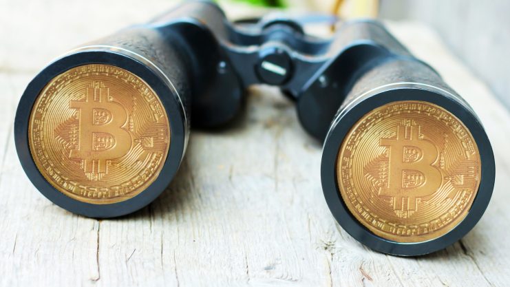 Ein Fernglas mit Bitcoin-Münzen auf den Linsen auf einer Holzoberfläche.