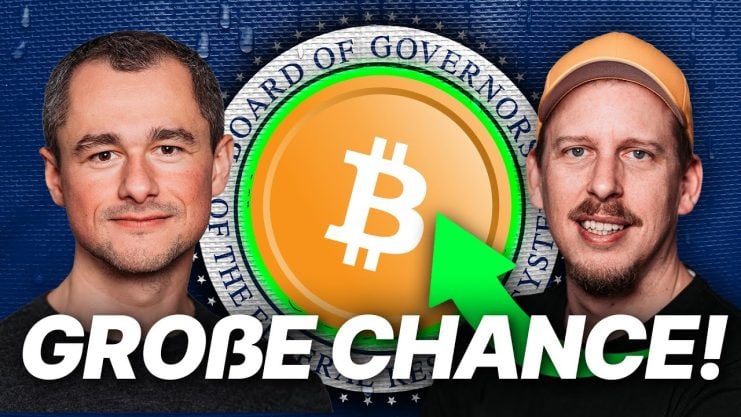 Zwei Männer stehen auf beiden Seiten eines großen Bitcoin-Symbols, auf das ein grüner Pfeil zeigt und darunter der deutsche Text „GROßE CHANCE!“ angezeigt wird, der auf mögliche Auswirkungen der US-Notenbank hinweist.