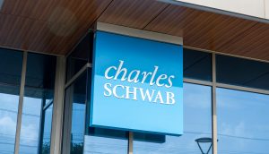 An der Außenseite eines modernen Glasgebäudes ist ein blaues Schild mit der weißen Aufschrift „Charles Schwab“ angebracht.