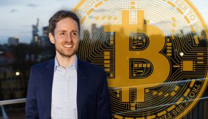 Dr. Jonas Groß posiert vor einem Bitcoin-Symbol, im Hintergrund ist die Frankfurter Skyline zu erkennen