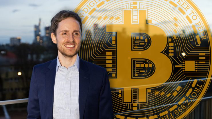 Dr. Jonas Groß posiert vor einem Bitcoin-Symbol, im Hintergrund ist die Frankfurter Skyline zu erkennen