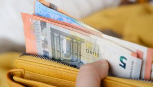 Eine Hand hält eine gelbe Brieftasche mit mehreren Euro-Banknoten und einer Karte mit einem teilweise sichtbaren Bitcoin-Verweis im offenen Reißverschlussfach.