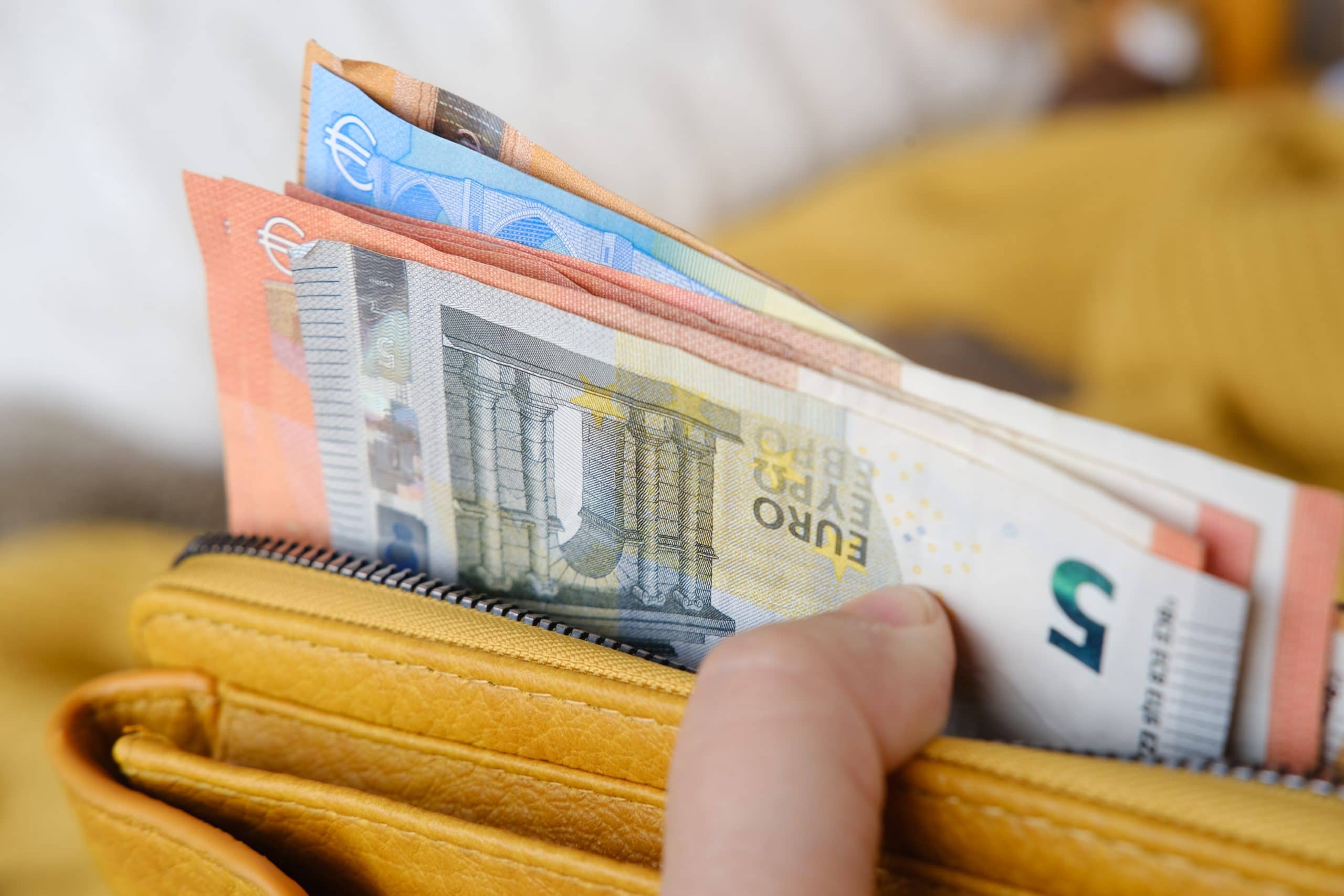 Eine Hand hält eine gelbe Brieftasche mit mehreren Euro-Banknoten und einer Karte mit einem teilweise sichtbaren Bitcoin-Verweis im offenen Reißverschlussfach.