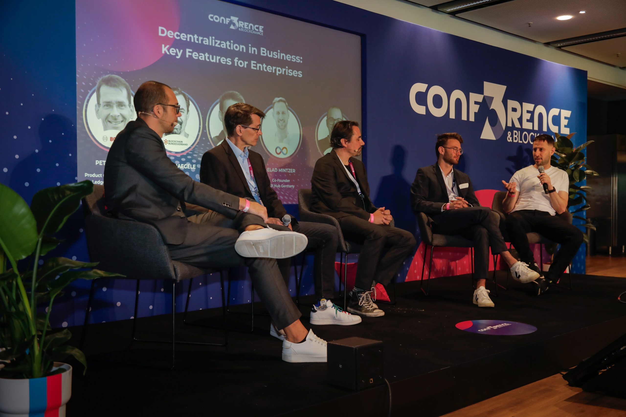 Fünf Männer sitzen auf einer Podiumsbühne der CONF3RENCE und diskutieren über Dezentralisierung in Unternehmen. Hinter ihnen zeigt ein Bildschirm das Thema und Fotos der Redner.