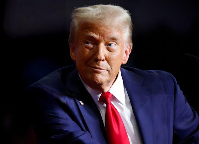 Donald Trump in einem dunkelblauen Anzug und einer roten Krawatte sitzt vor einem dunklen Hintergrund.