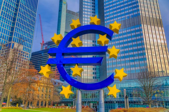 An einem klaren Tag steht vor modernen Wolkenkratzern und einem Kran eine Skulptur des Eurosymbols, umgeben von gelben Sternen, und symbolisiert Widerstandsfähigkeit angesichts der Inflationssorgen.