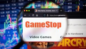Eine Lupe fokussiert auf die GameStop-Website, die auf einem Computerbildschirm angezeigt wird, vor dem Hintergrund dynamischer Börsendiagramme und -charts.