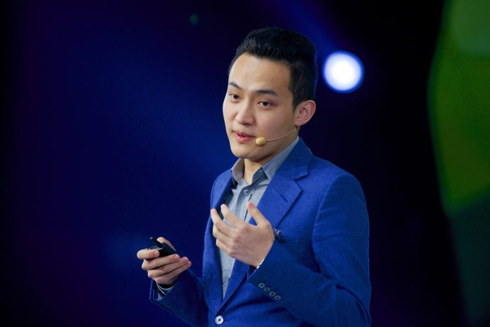 Justin Sun ist elegant in einen blauen Anzug gekleidet, spricht selbstbewusst in ein Headset-Mikrofon und leitet das Publikum auf der schwach beleuchteten Bühne nahtlos mit seinem Klicker an.