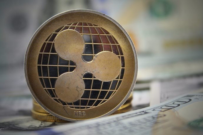 Die Kryptowährung Ripple steht im Mittelpunkt. Ihr vom Globus inspiriertes Logo ist auf einer Oberfläche mit verschwommenen US-Dollarscheinen im Hintergrund zu sehen, während nach einer kürzlichen Ankündigung von Coinbase die Spannung über XRP-Futures steigt.