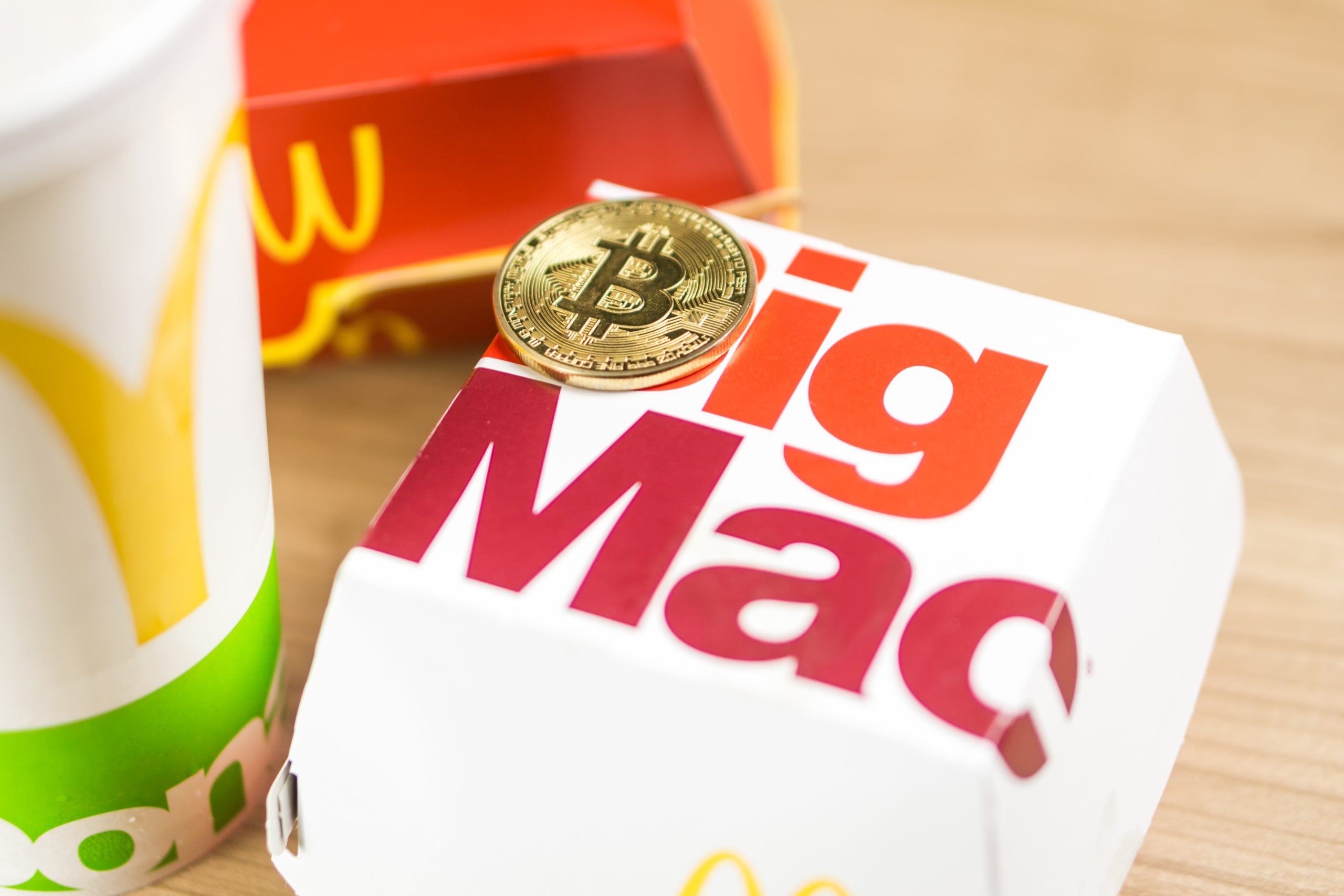 Ein glänzender Bitcoin liegt auf einer Big-Mac-Schachtel, im Hintergrund lugt ein Hauch des typischen McDonald's-Getränkebechers und der Pommes-Schachtel hervor, wodurch die Welt der digitalen Währung perfekt mit Fast-Food-Nostalgie verschmilzt.