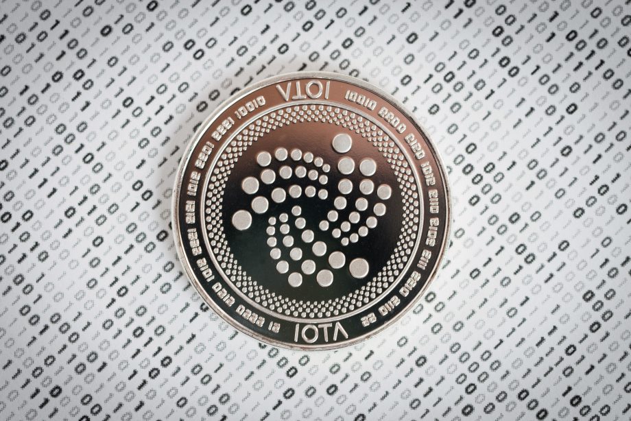 Eine silberne IOTA-Kryptowährungsmünze wird auf einem gemusterten Hintergrund mit Binärcodesymbolen angezeigt.