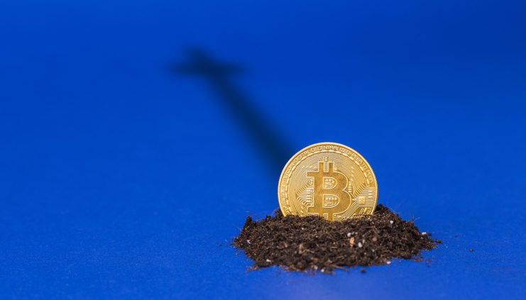 Eine teilweise im Boden vergrabene goldene Bitcoin-Münze auf blauem Hintergrund, deren Schatten die Form eines Todeskreuzes bildet.