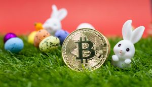 Eine goldene Bitcoin-Münze liegt auf grünem Gras, umgeben von kleinen dekorativen Ostereiern und zwei weißen Hasenfiguren vor einem leuchtend roten Hintergrund.