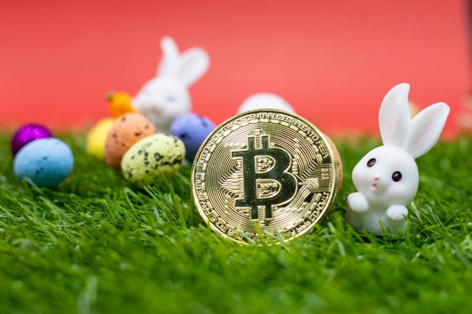 Eine goldene Bitcoin-Münze liegt auf grünem Gras, umgeben von kleinen dekorativen Ostereiern und zwei weißen Hasenfiguren vor einem leuchtend roten Hintergrund.