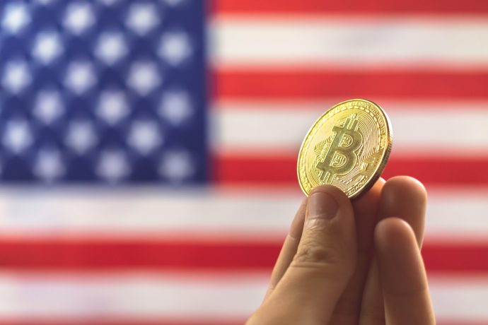 Eine Hand hält eine goldfarbene Bitcoin-Reserve-Münze vor einer verschwommenen US-Flagge, die den wachsenden Einfluss der Kryptowährung in Amerika symbolisiert, der sogar Orte wie Oklahoma erreicht.