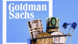 Vor einem Bildschirm mit dem Logo von Goldman Sachs wird ein Miniatur-Einkaufswagen mit gerollten US-Dollar- und Euro-Scheinen sowie Kryptowährungsmünzen präsentiert.