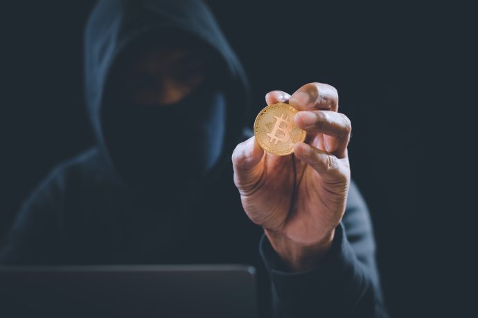Eine mysteriöse Gestalt in einem dunklen Kapuzenpullover und mit Hacker-ähnlichem Auftreten hält eine Bitcoin-Münze hoch, die vom Schein ihres Laptops vor dem schattigen Hintergrund beleuchtet wird.