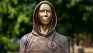 Eine Bronzestatue einer vermummten Figur, die Satoshi Nakamoto symbolisiert, zeigt stolz ein Bitcoin-Logo auf der Brust, vor einem beruhigenden, verschwommenen Hintergrund aus grünen Blättern.