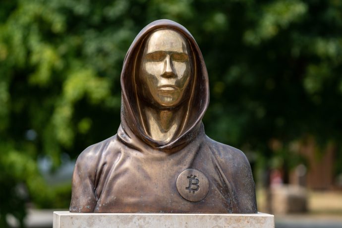 Eine Bronzestatue einer vermummten Figur, die Satoshi Nakamoto symbolisiert, zeigt stolz ein Bitcoin-Logo auf der Brust, vor einem beruhigenden, verschwommenen Hintergrund aus grünen Blättern.