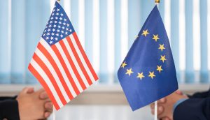 Zwei kleine Tischfahnen, eine der Vereinigten Staaten und eine der EU, werden zwischen zwei einander gegenübersitzenden Personen platziert und symbolisieren eine Diskussion über regulatorische Klarheit.