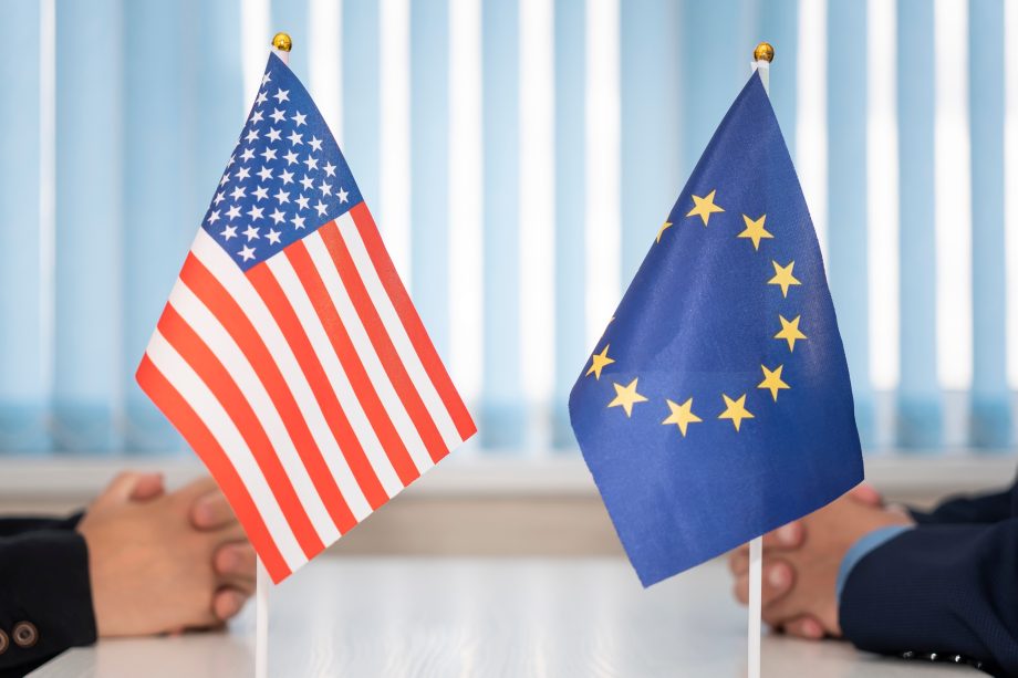Zwei kleine Tischfahnen, eine der Vereinigten Staaten und eine der EU, werden zwischen zwei einander gegenübersitzenden Personen platziert und symbolisieren eine Diskussion über regulatorische Klarheit.