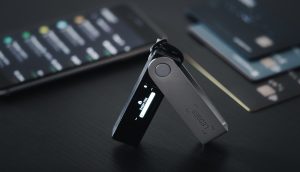 Auf einer dunklen Oberfläche ist eine Hardware-Kryptowährungs-Wallet von Ledger geöffnet und aktiv. Auf einem Smartphone in der Nähe wird die Ledger-App angezeigt und im Hintergrund sind zwei Zahlungskarten zu sehen.