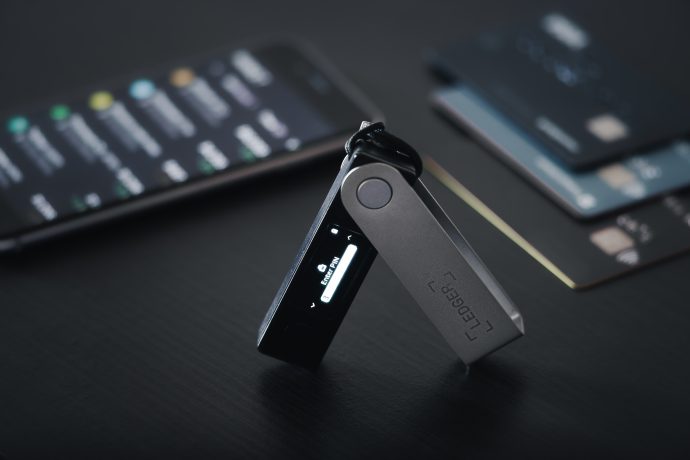 Auf einer dunklen Oberfläche ist eine Hardware-Kryptowährungs-Wallet von Ledger geöffnet und aktiv. Auf einem Smartphone in der Nähe wird die Ledger-App angezeigt und im Hintergrund sind zwei Zahlungskarten zu sehen.