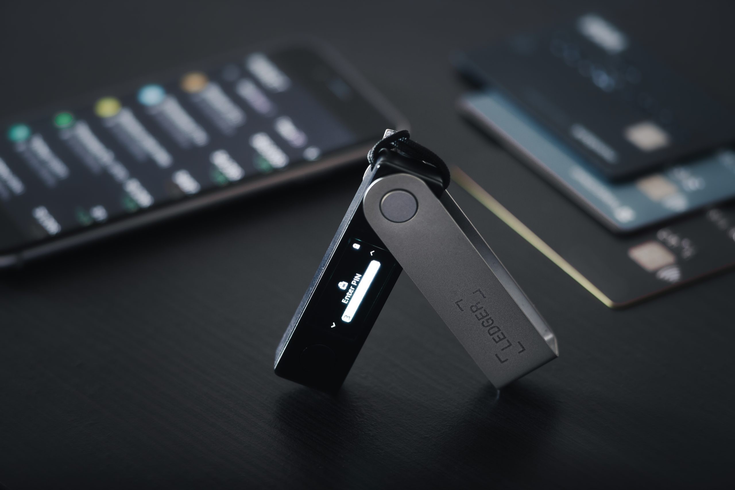 Auf einer dunklen Oberfläche ist eine Hardware-Kryptowährungs-Wallet von Ledger geöffnet und aktiv. Auf einem Smartphone in der Nähe wird die Ledger-App angezeigt und im Hintergrund sind zwei Zahlungskarten zu sehen.